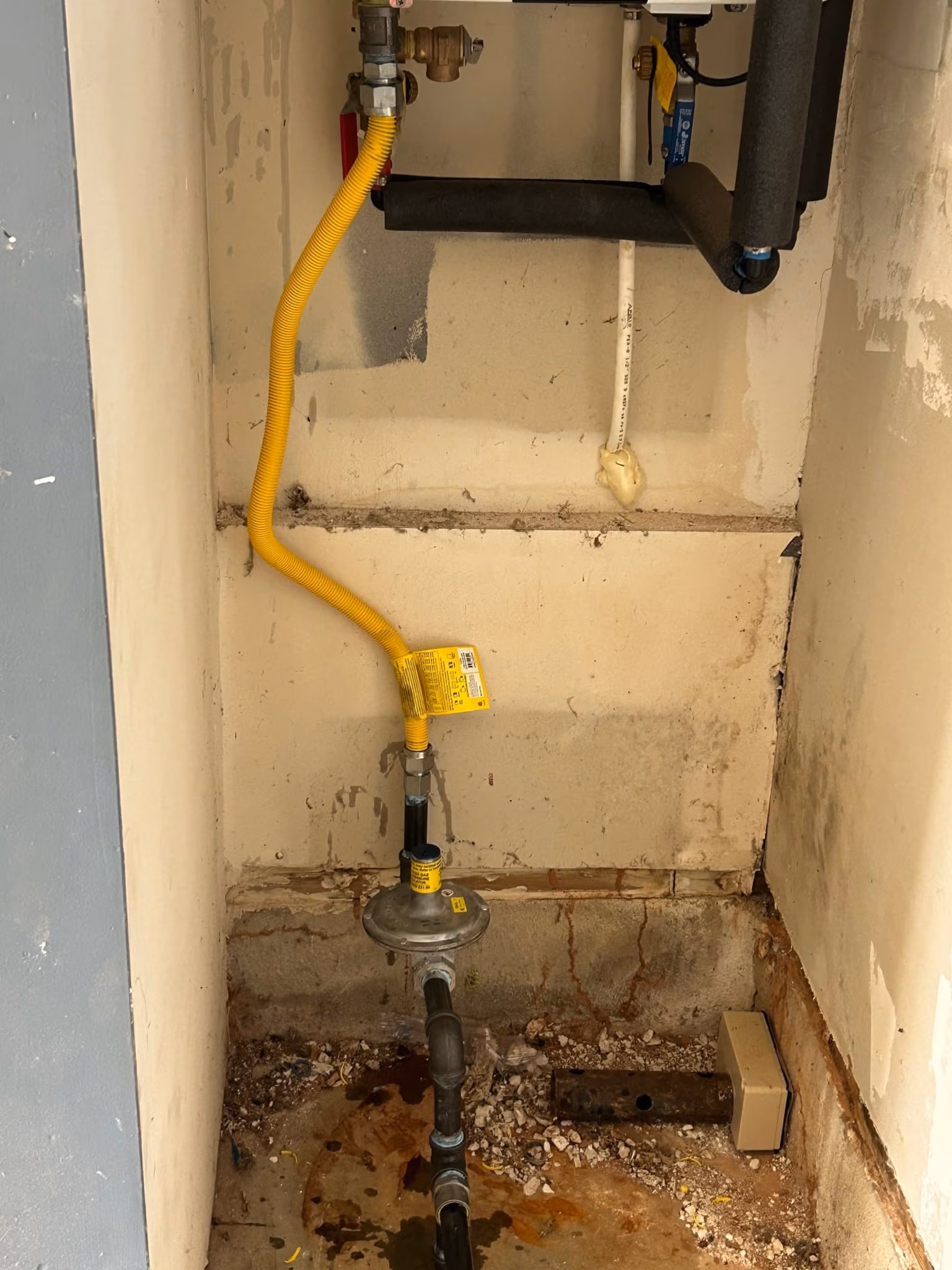 Yellow flexible gas line connected to a valve and pipe in a dirty, unfinished utility closet.
