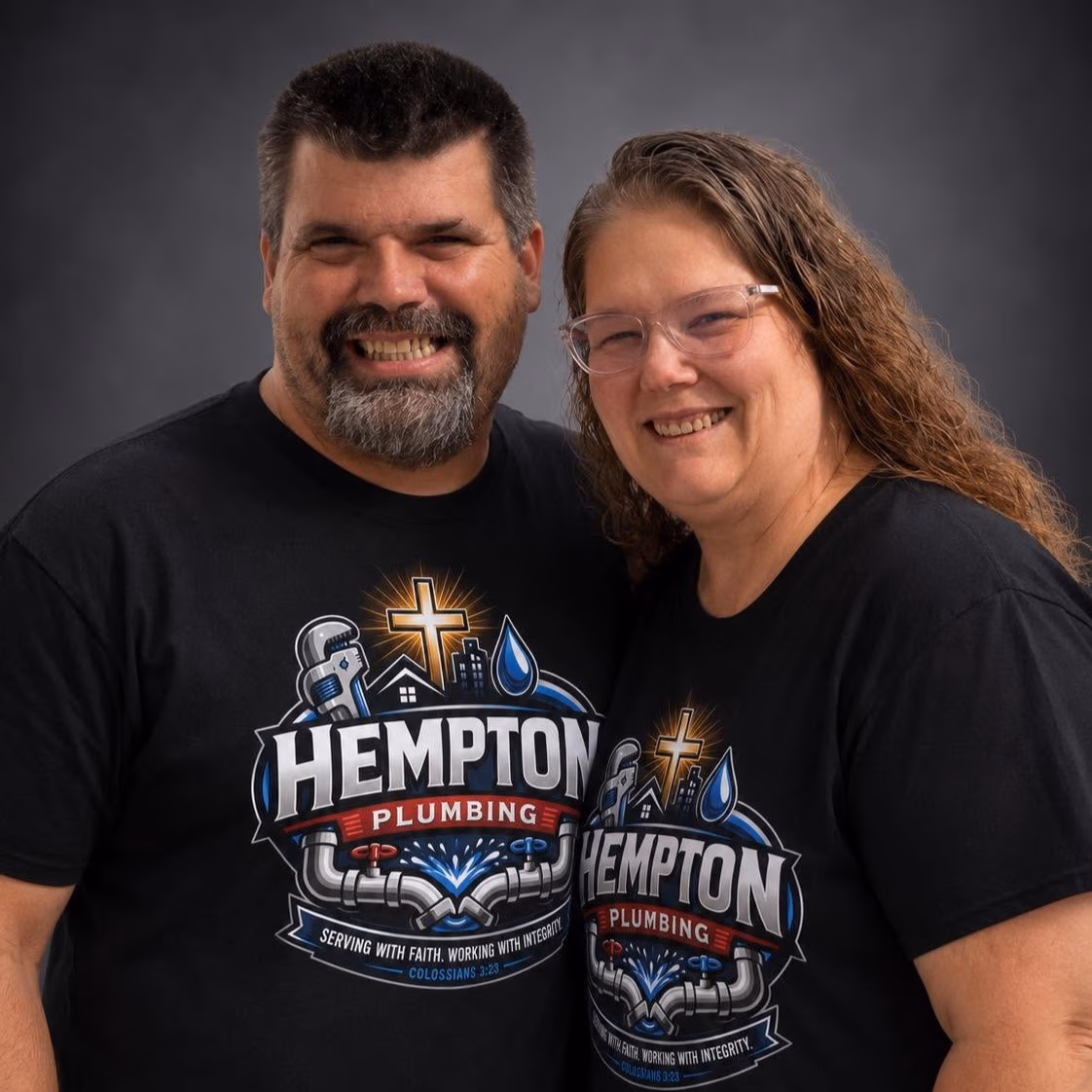 Smiling man and woman wearing matching Hempton Plumbing T-shirts with cross and pipes logo.