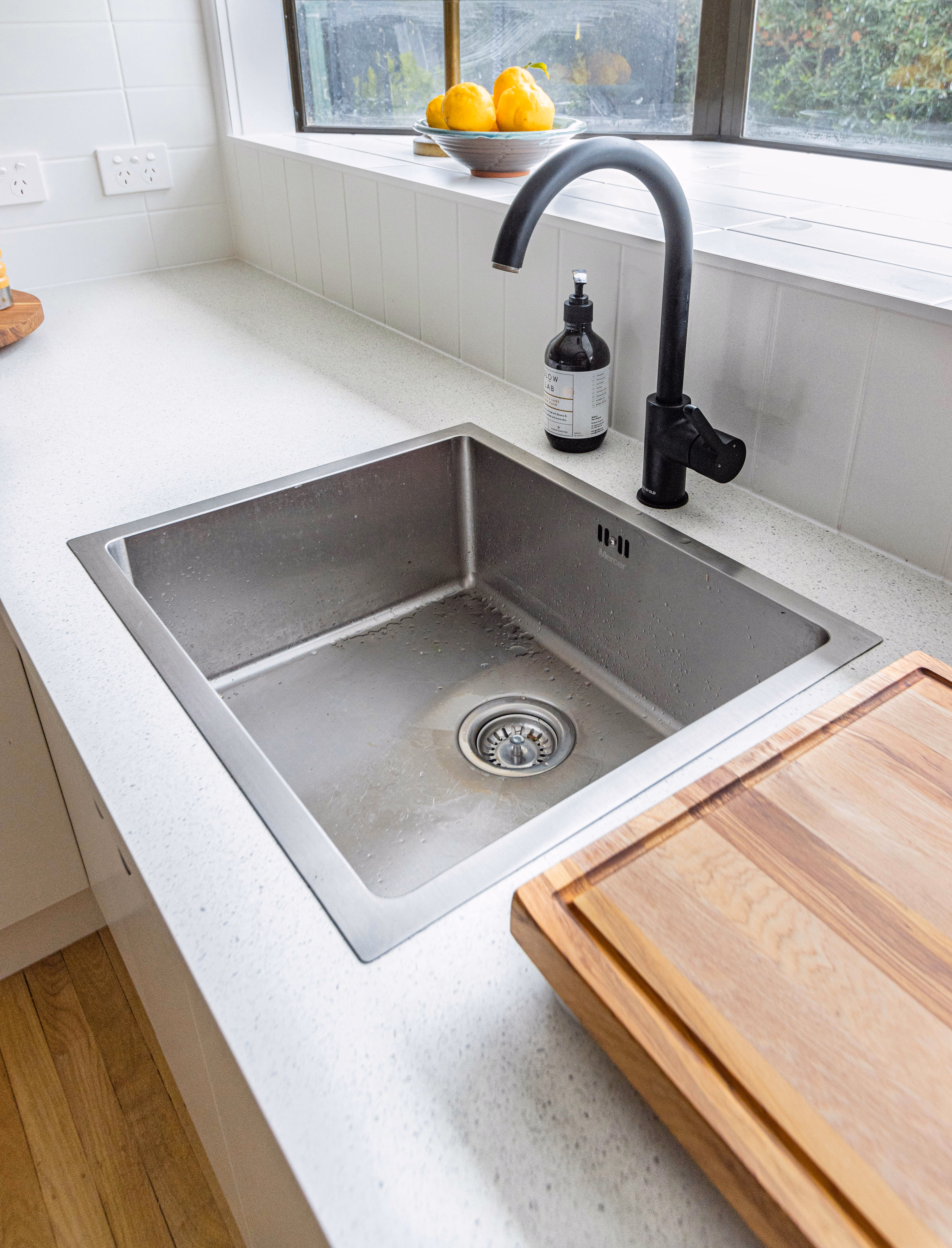 Stainless steel kitchen sink with black faucet, soap dispenser, wooden cutting board, and lemons in a bowl.
