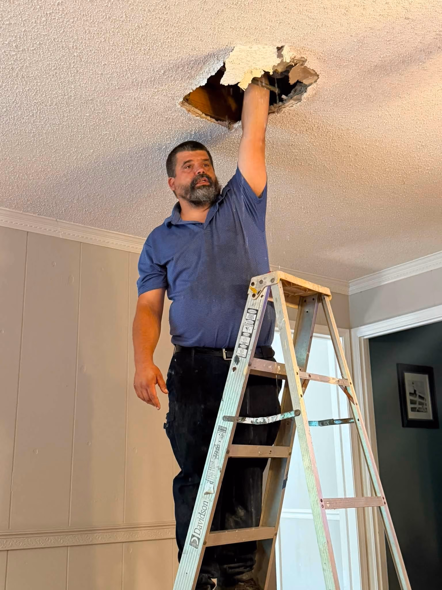 Man on a ladder reaching into a hole in the ceiling for repair work.