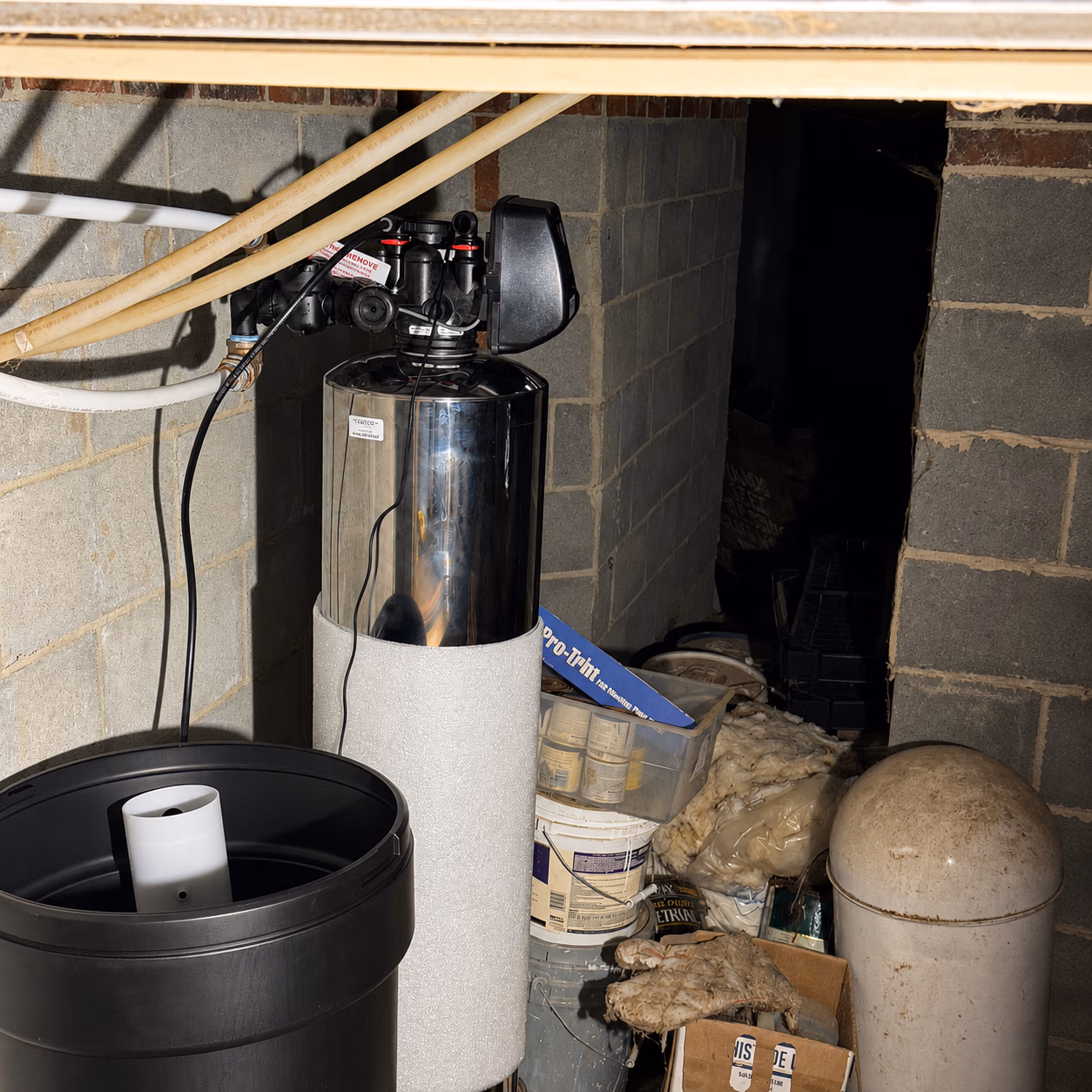 Water softener system and plastic containers in a basement with cinder block walls and clutter.